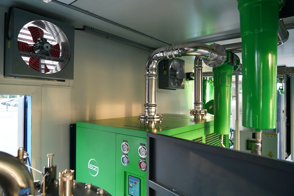 Inside a skid mounted air compressor station showing green compressor, stainless steel piping, isolation valves and ventilation fans